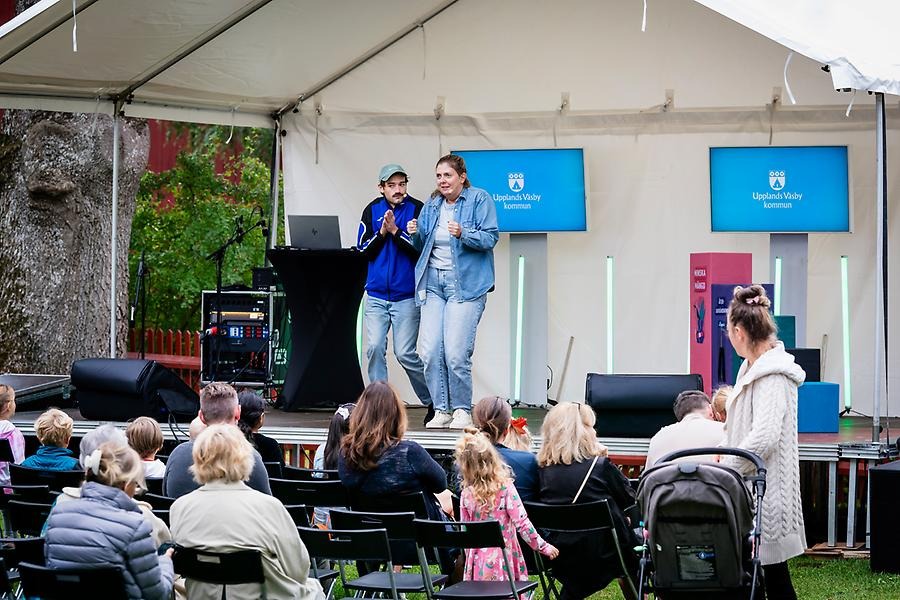 Två dramapedagoger, en man och en kvinna, från Teater & Dramaskolan uppträder på scen under Återbruksfestivalen i september. Nedanför scenen syns en publik i blandade åldrar som tittar på föreställningen